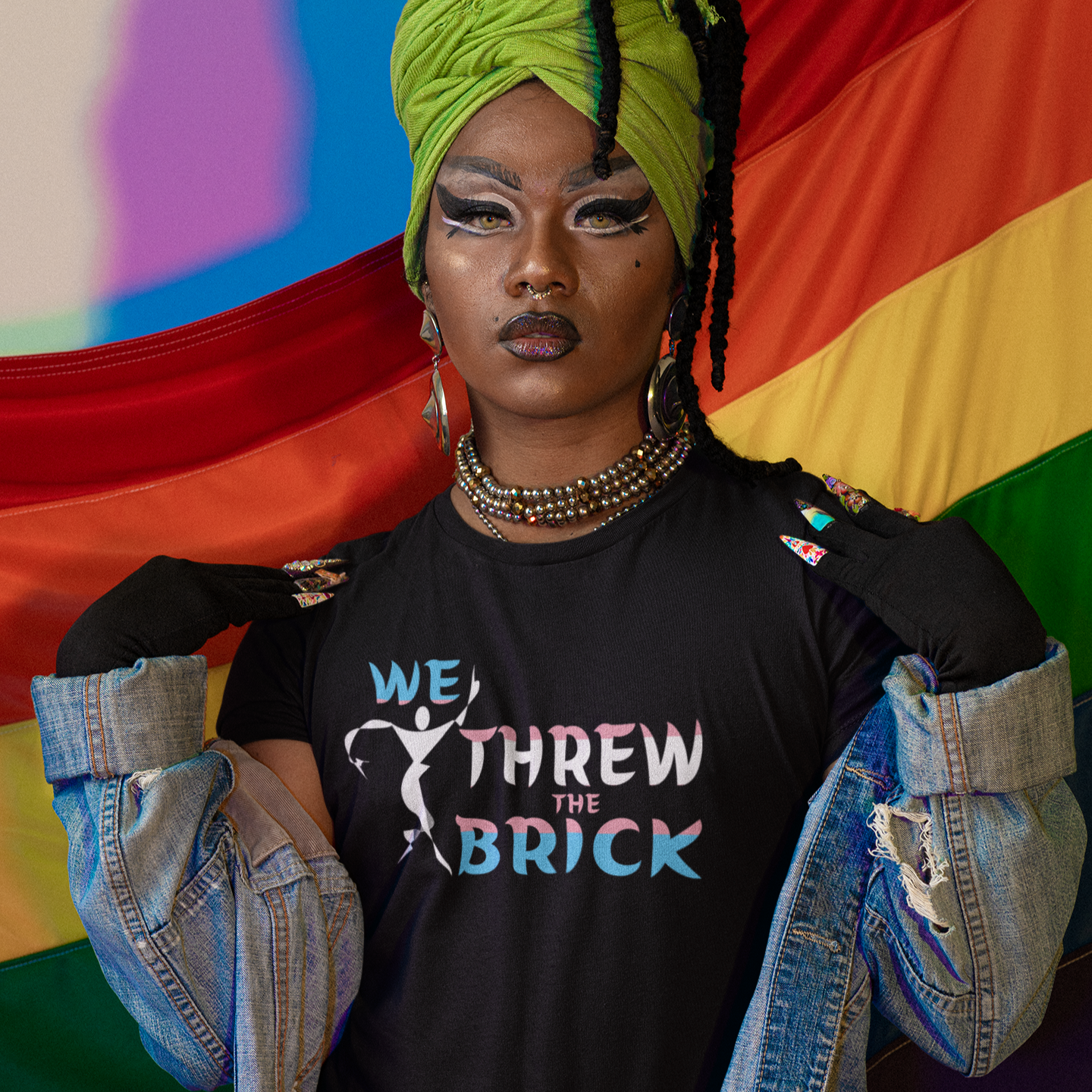 Person wearing a black t-shirt with 'We Threw the Brick' design, standing in front of a rainbow flag.